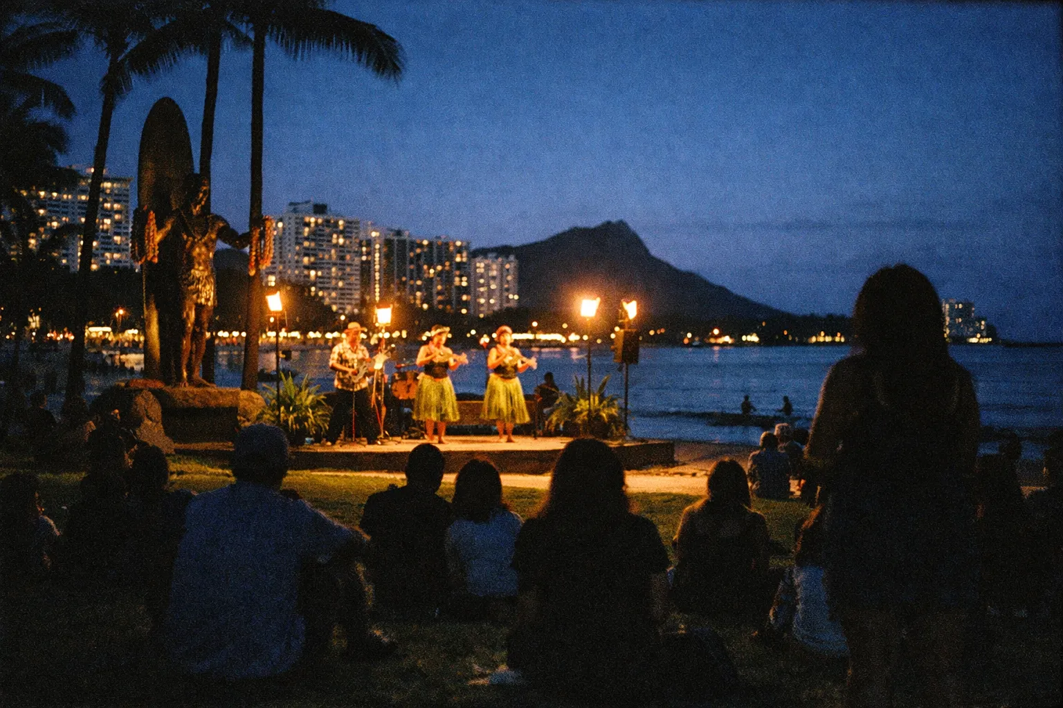 A crowd watches a hula performance near the Duke Kahanamoku statue on Waikiki Beach at dusk with Diamond Head in view.