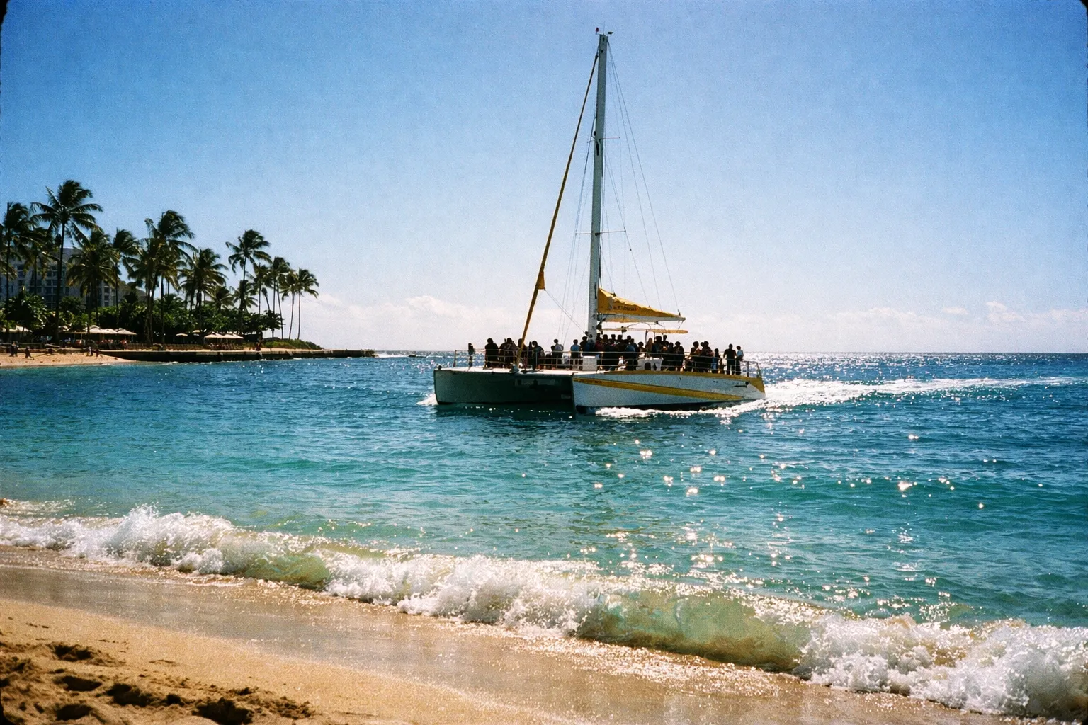 A catamaran with passengers sails near Waikiki Beach with palm trees lining the shore.