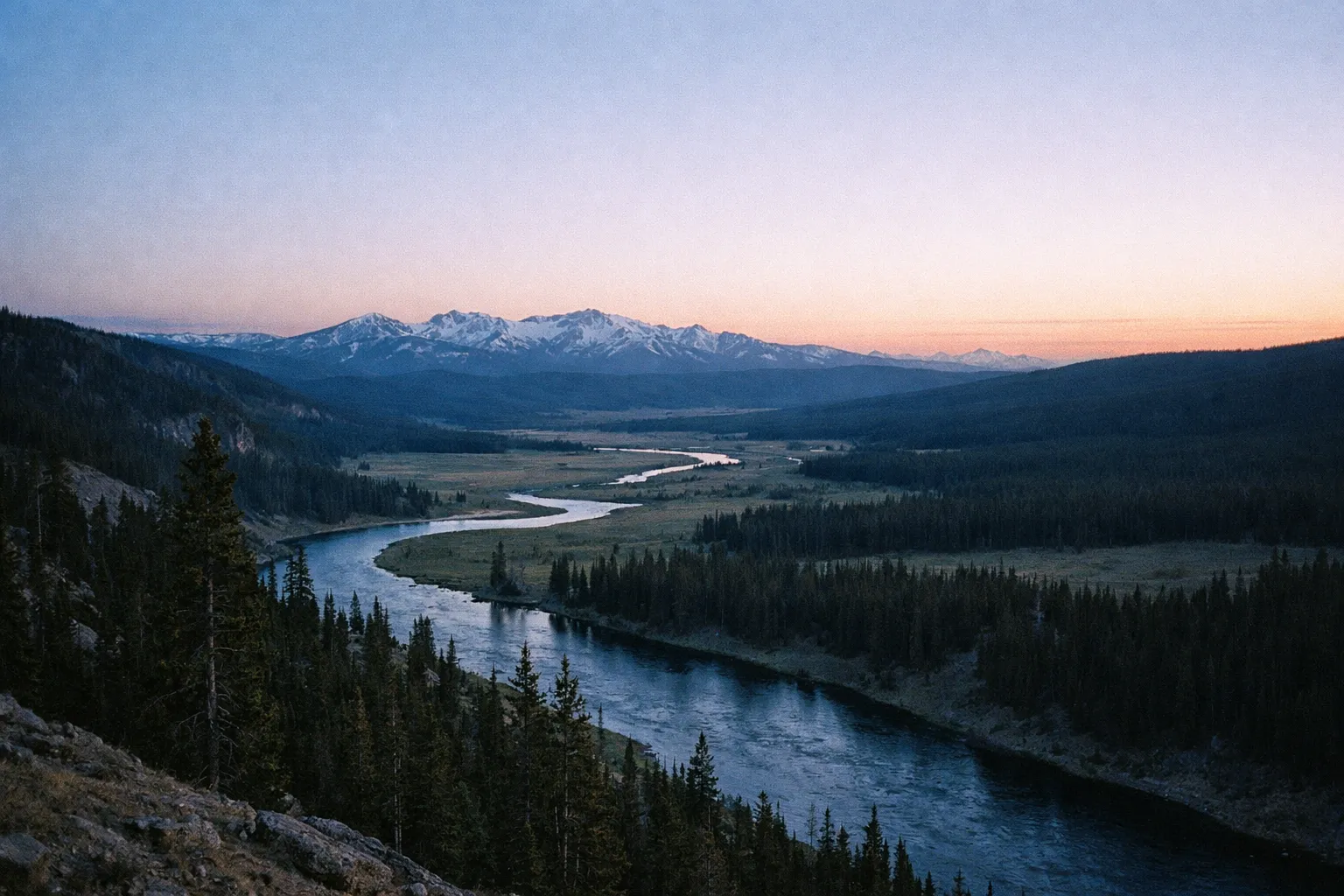 A winding river flows through a forested valley with snow-capped mountains in Yellowstone at sunset.