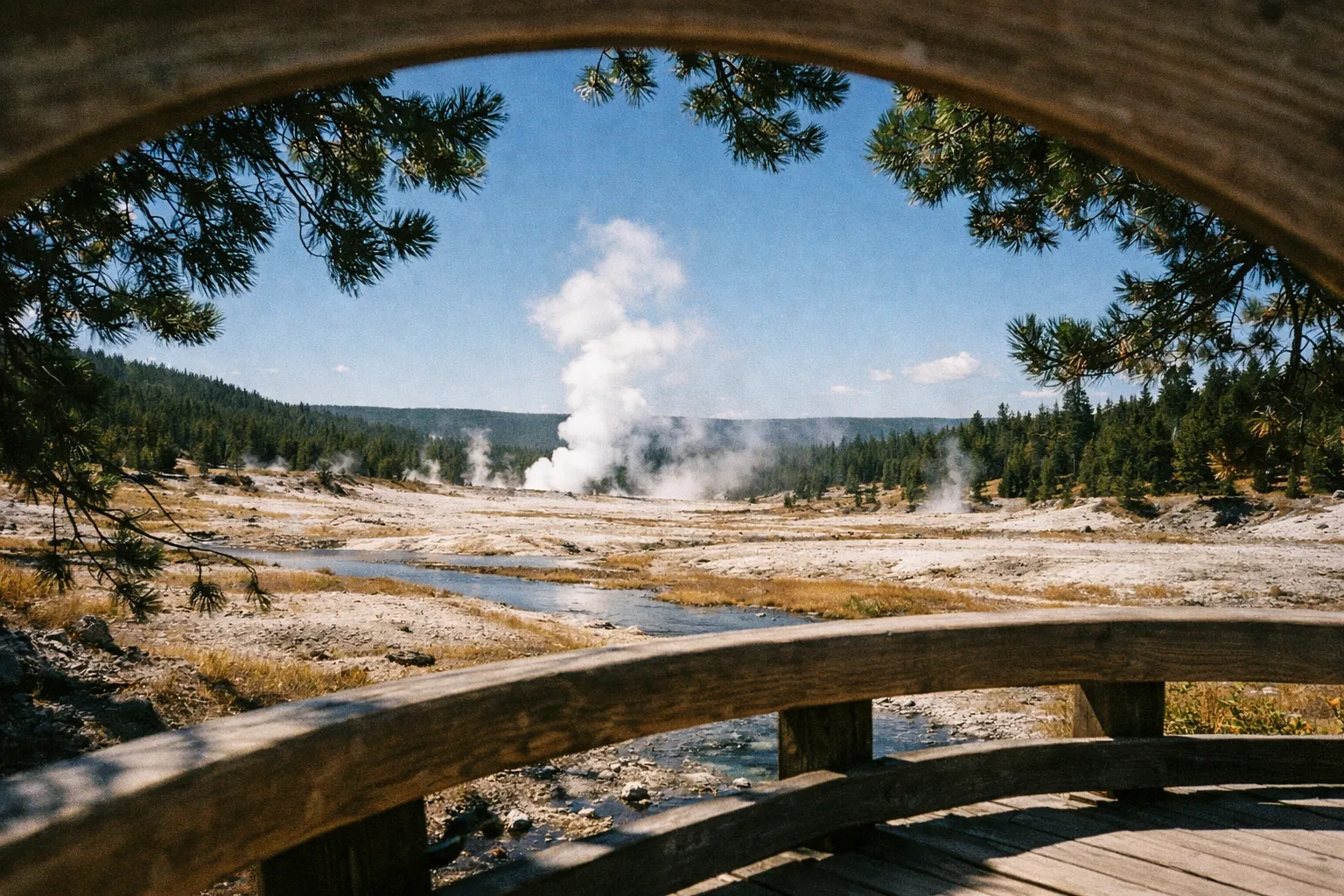 A geyser steams in a wide open landscape, viewed from behind a wooden railing in Yellowstone National Park.