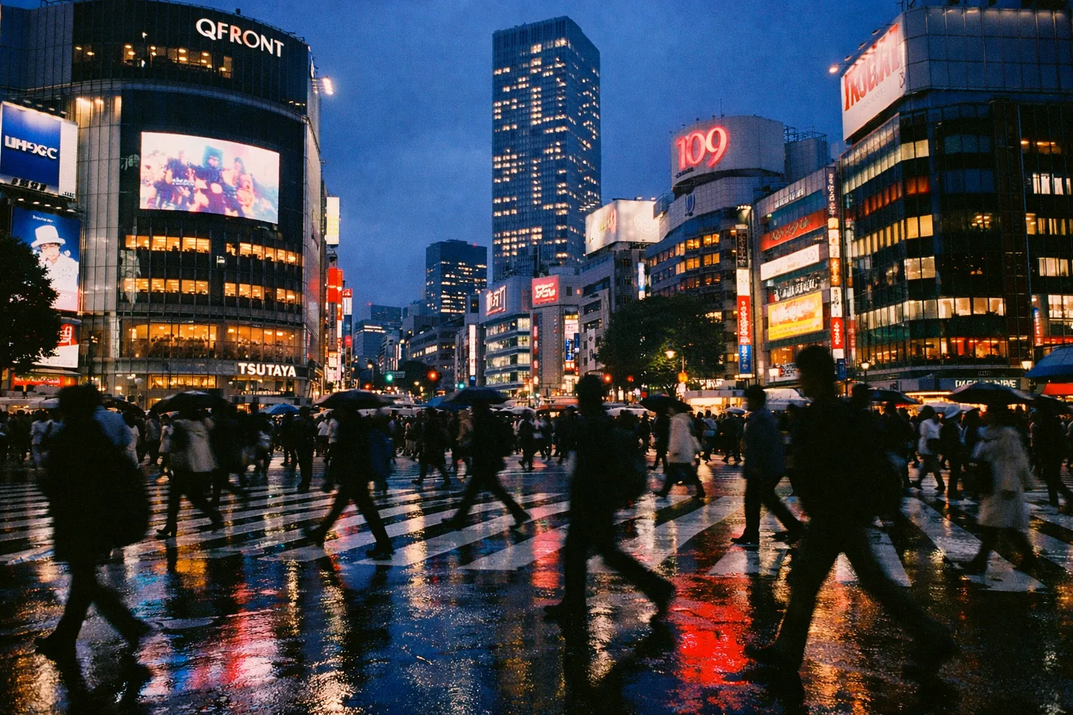 Crowds with umbrellas cross Shibuya Crossing in Tokyo at night, surrounded by illuminated buildings and signs.
