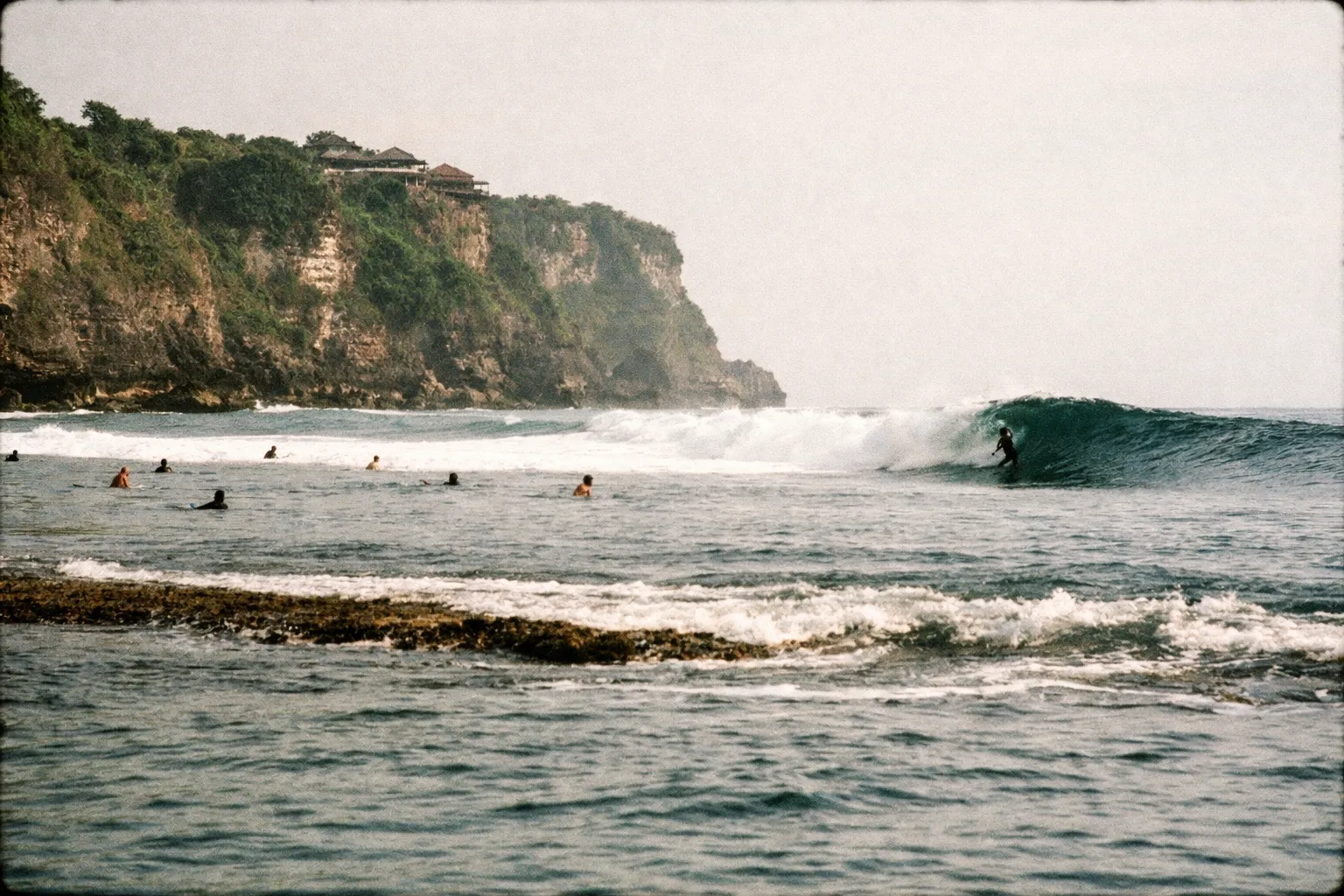 Surfers wait on their boards and ride waves near dramatic cliffs at Uluwatu Beach in Bali.