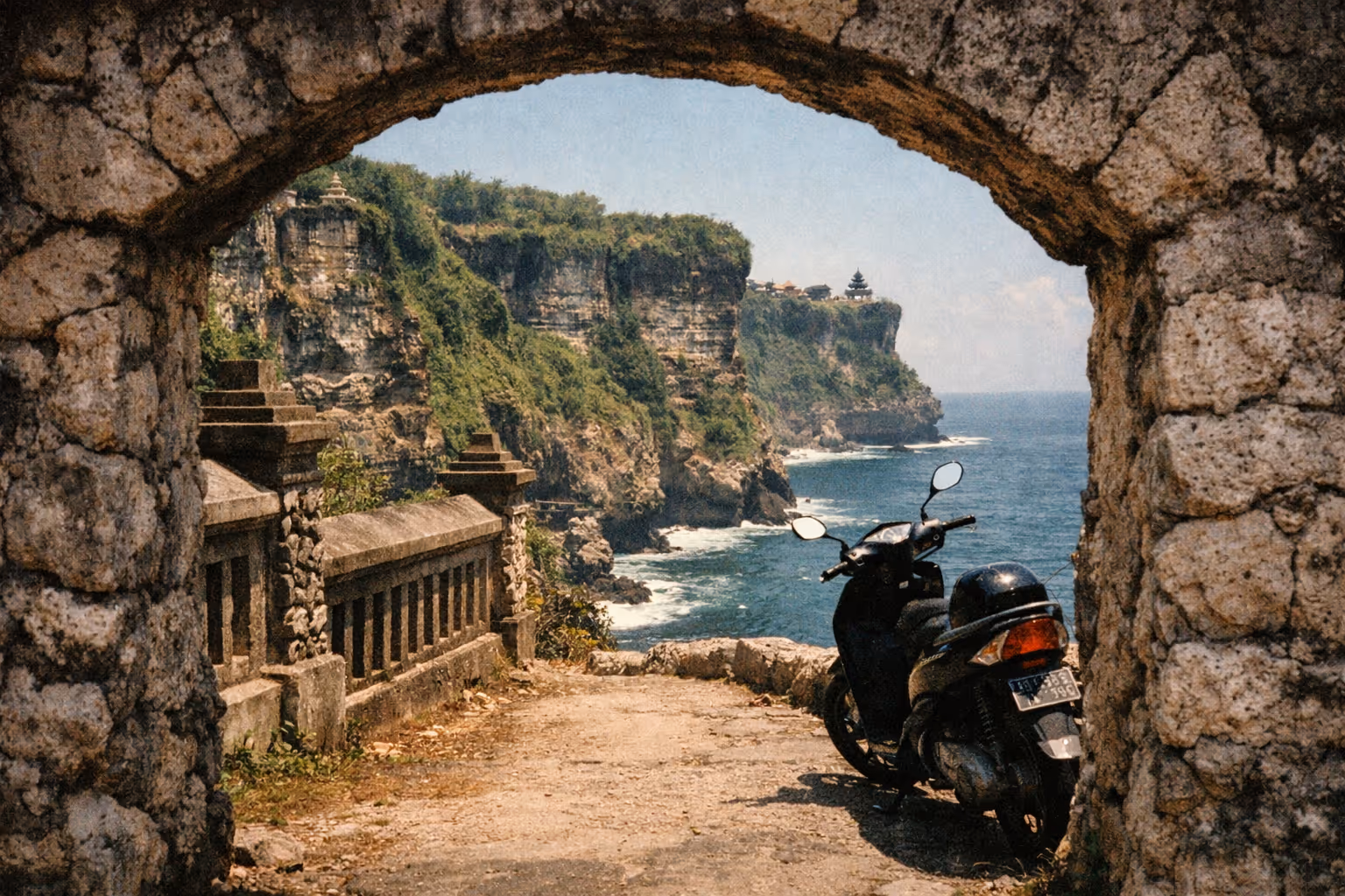 A scooter is parked under a stone archway overlooking Uluwatu Temple on dramatic Bali sea cliffs.