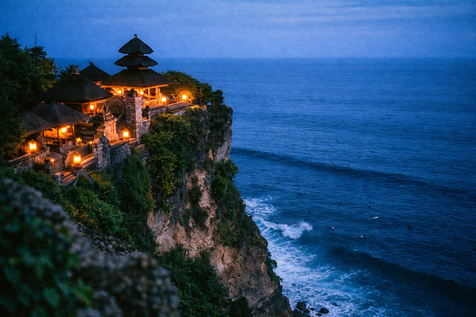 Uluwatu Temple perched on a cliff above the ocean with surfers visible in the waves below.