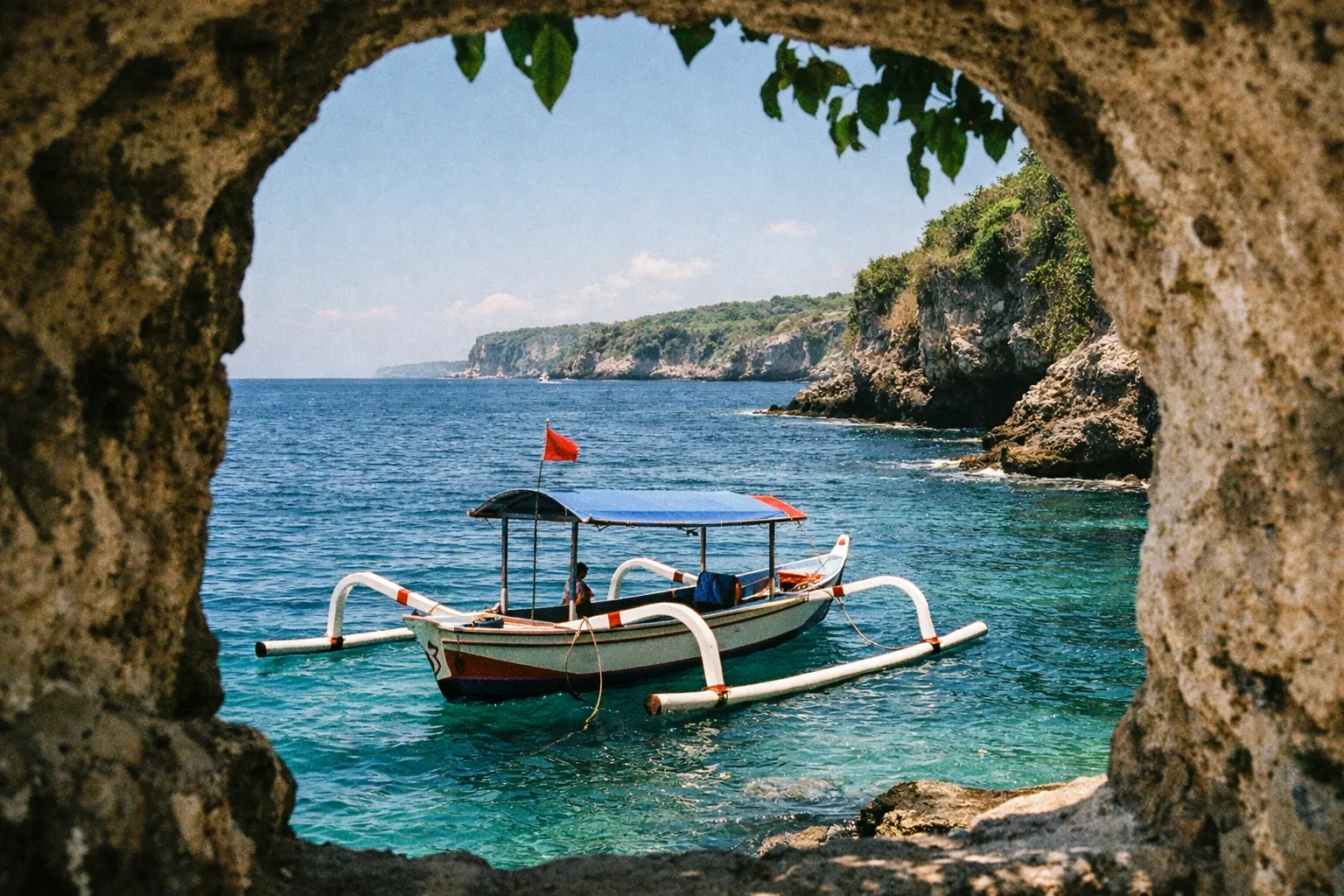 A traditional Balinese fishing boat floats near rocky cliffs and clear blue water on the coastline.