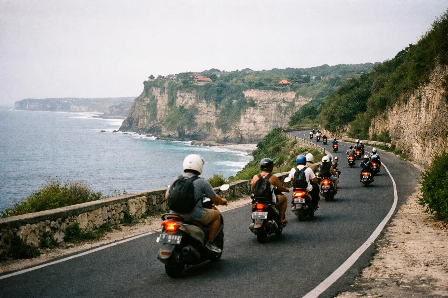 People ride scooters along a coastal road with dramatic cliffs and ocean views near Uluwatu, Bali.