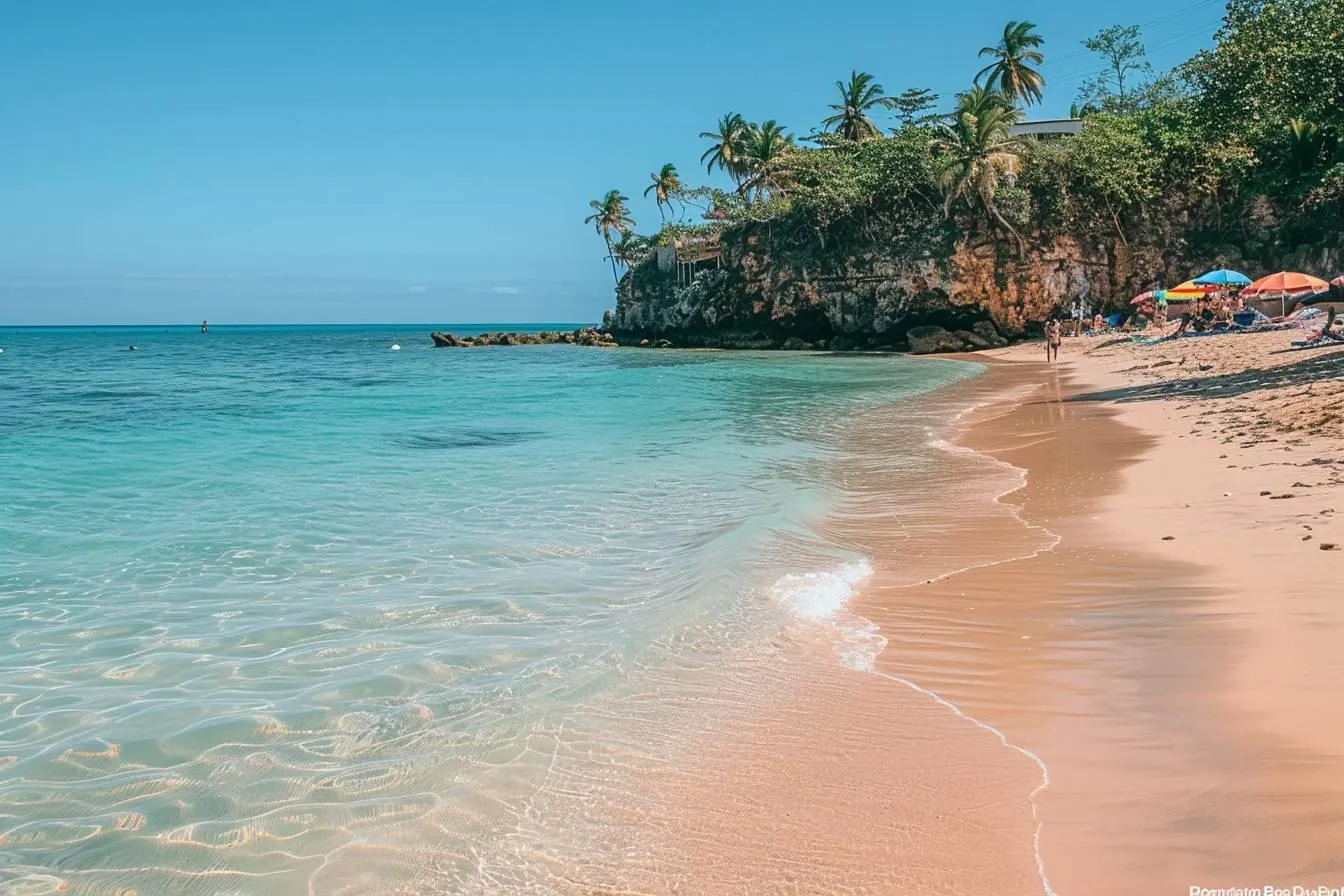 People relax under umbrellas on a sandy beach with turquoise water and palm trees along a rocky cliff.