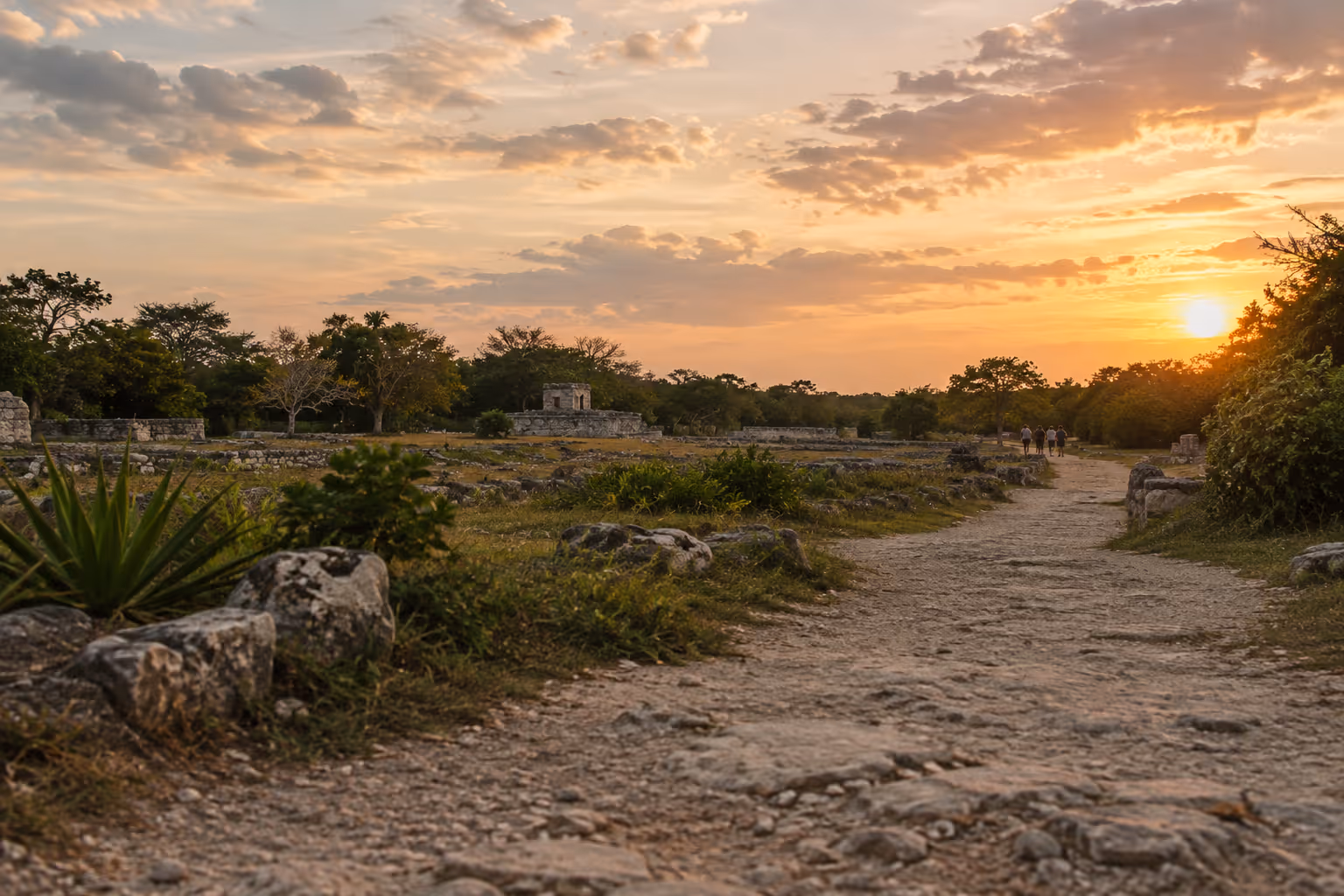 Visitors walk a stone path through low Maya ruins beneath a sunset sky.