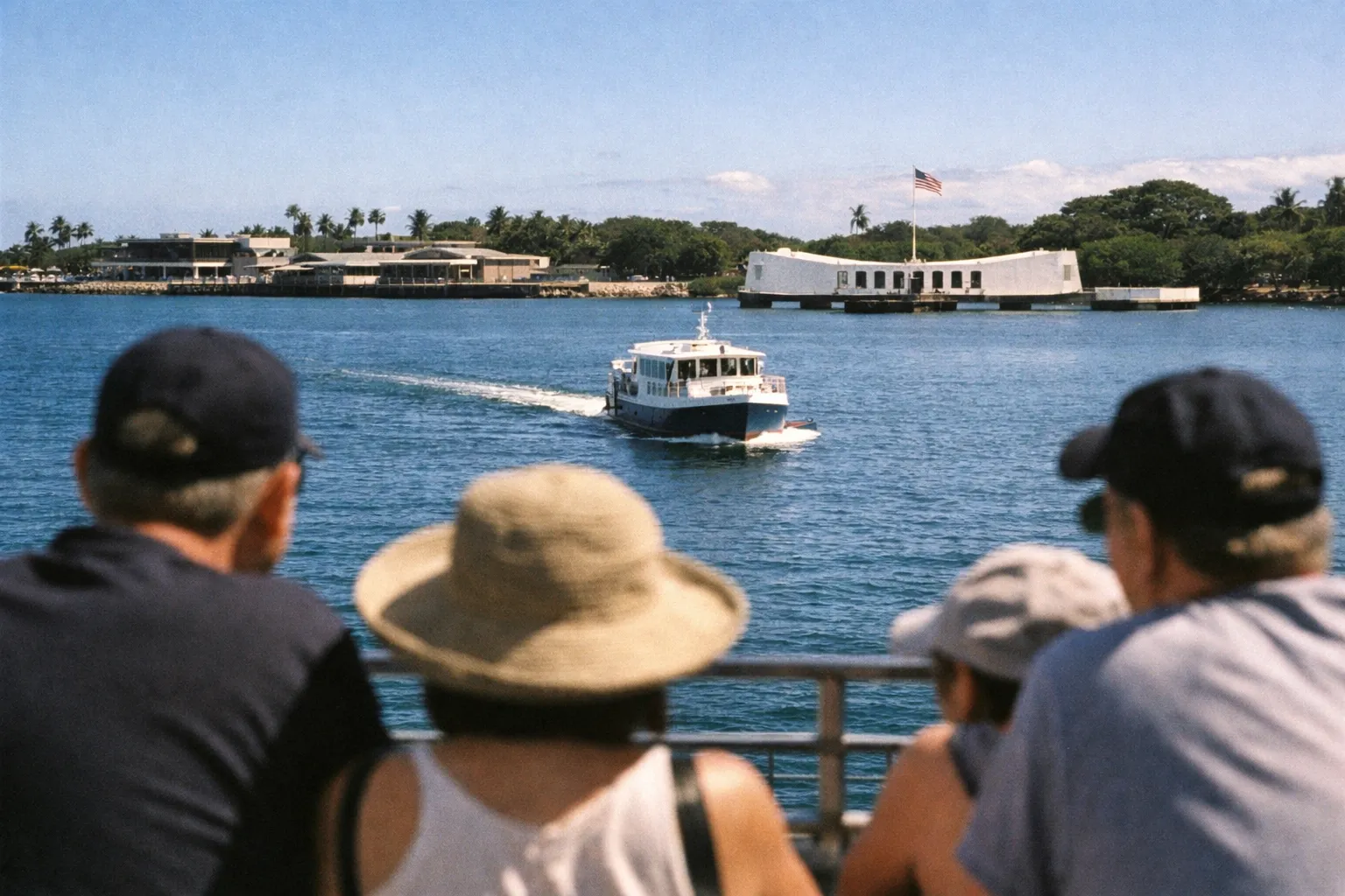 Visitors watch a boat approach the USS Arizona Memorial at Pearl Harbor under a clear blue sky.