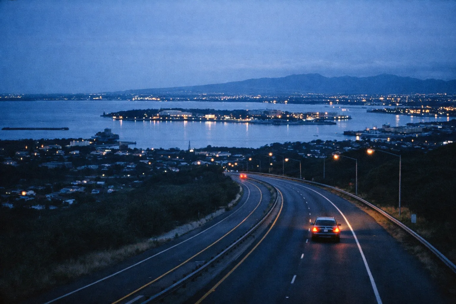 A car drives on a curving highway overlooking Pearl Harbor at dusk, with city lights reflecting on the water.