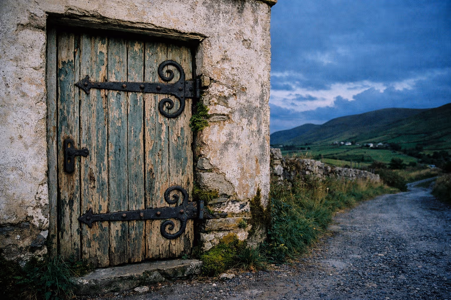 Old wooden door with ornate hinges on a weathered stone building next to a rural dirt road and hills